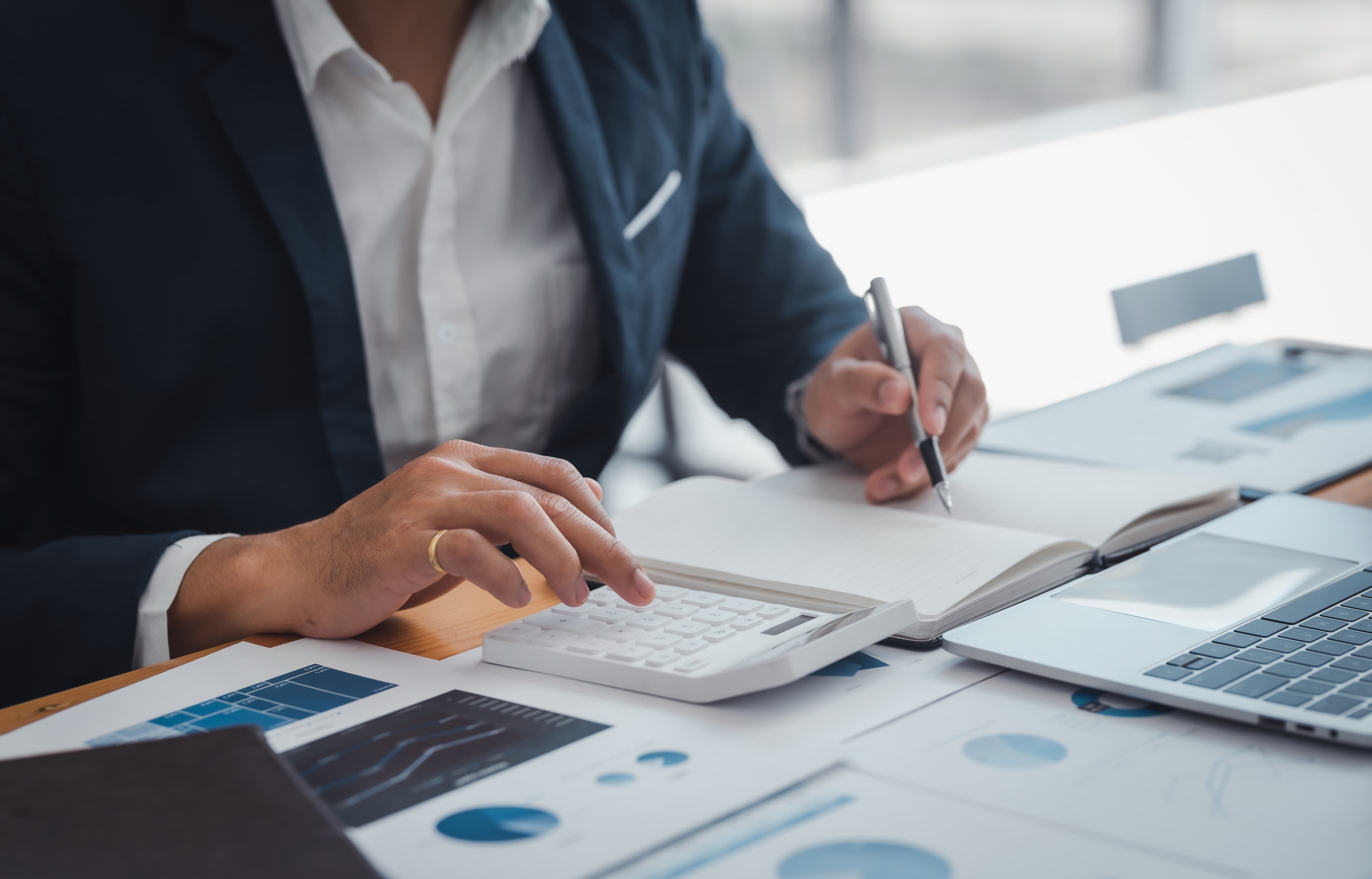 A person in a suit calculating with a notebook and pen, surrounded by charts and a laptop on a wooden desk, conveying a focused and professional tone.