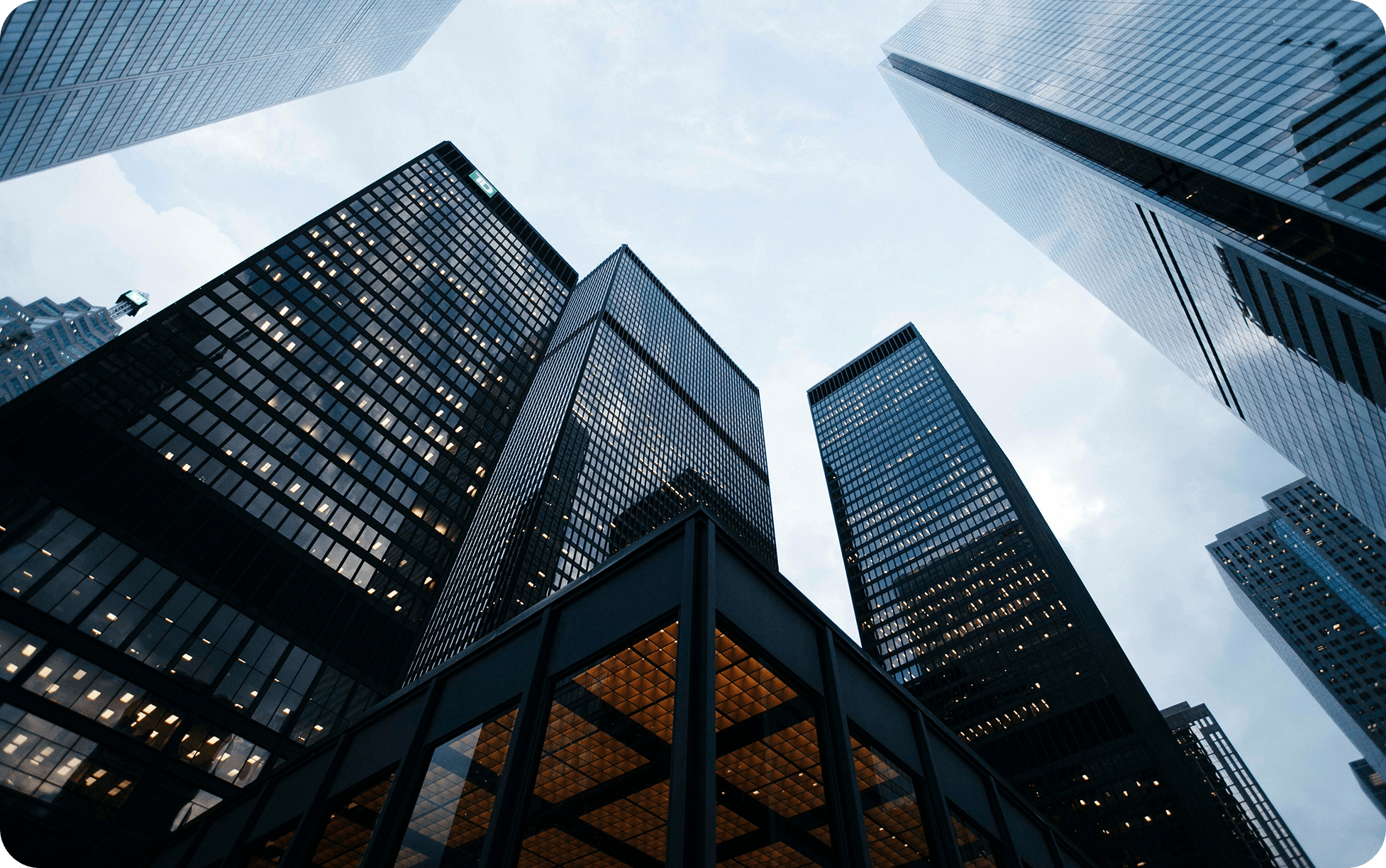 Skyscrapers rise against a cloudy sky, with their reflective glass facades gleaming. The perspective is upward, evoking grandeur and urban ambition.