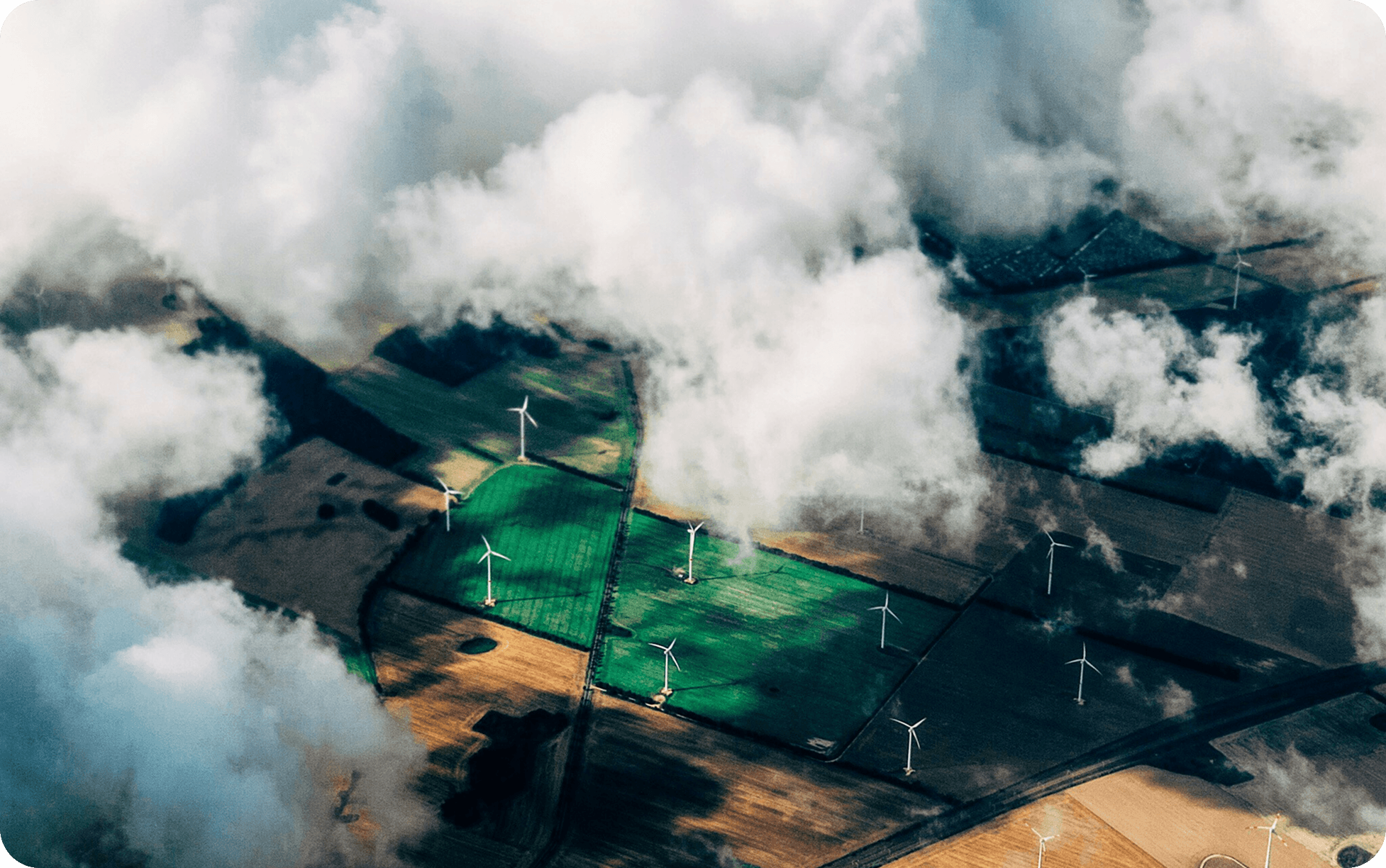 Aerial view showcasing a farm with fields and several wind turbines in the background against a clear blue sky.