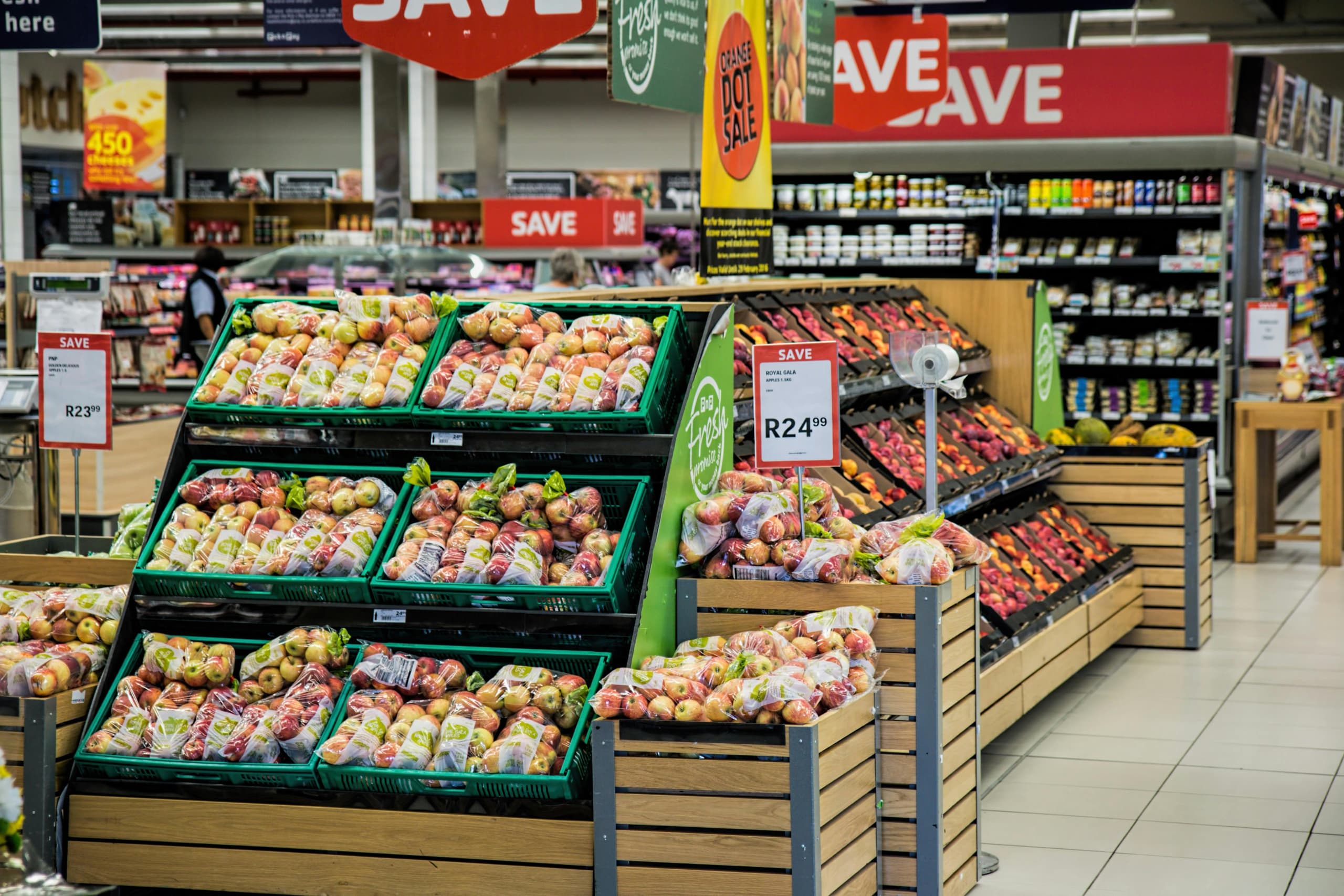 El pasillo de una tienda de comestibles muestra manzanas de colores vivos en estantes verdes, marcadas con precios de oferta, entre otros productos de colores.